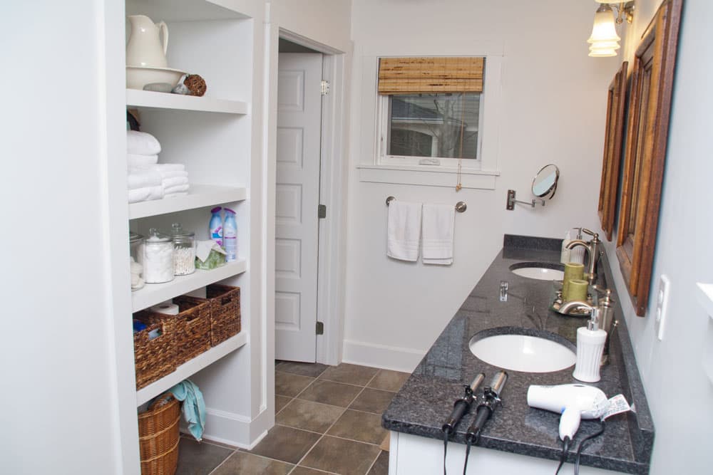 Renovated bathroom with dark granite double vanity, built-in white shelving, and bamboo window shade