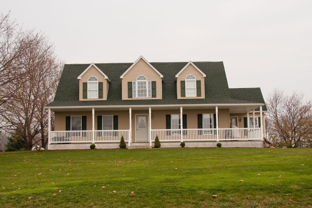 Two-story beige modular home with white wraparound porch and dark roof