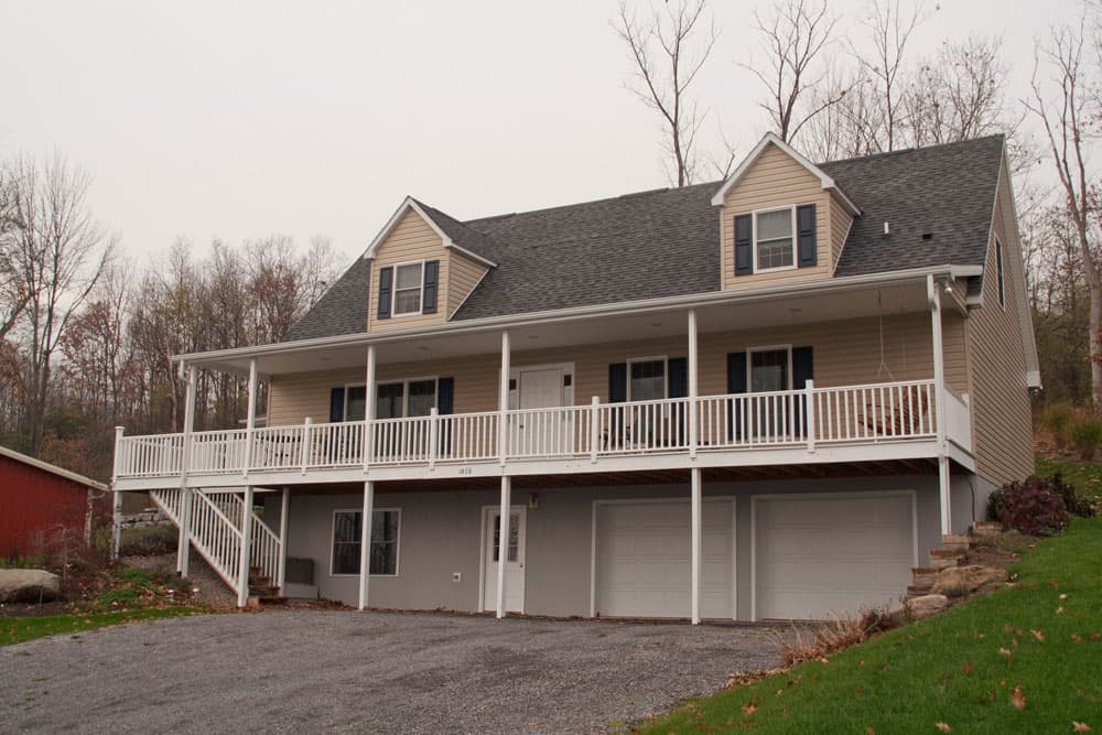 Tan modular home with white wrap-around porch and two-car garage below