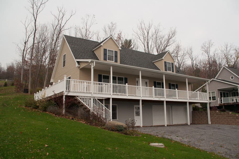 Two-story modular home with tan siding, wrap-around porch, and three-car garage