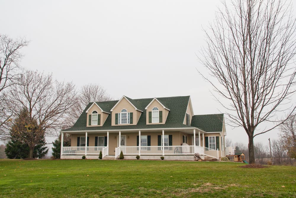 Two-story modular home with green roof, beige siding, and wraparound porch