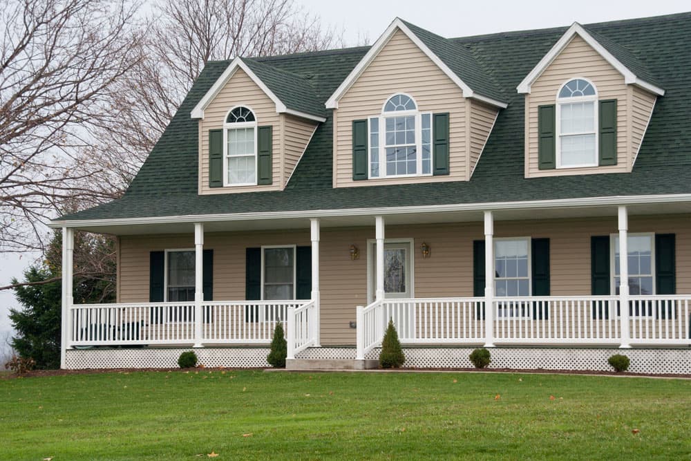 Beige two-story modular home with green shutters, three dormers, and white wraparound porch