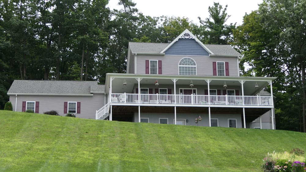Two-story modular home with vinyl siding, red shutters, and wraparound porch on hillside property