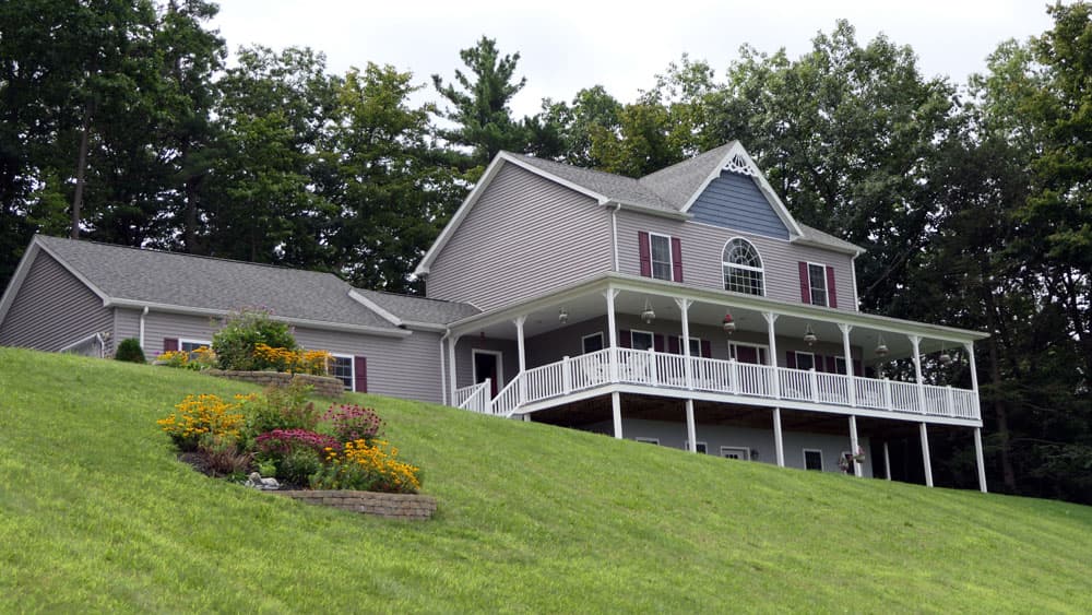 Gray modular home with white wraparound porch on landscaped hillside